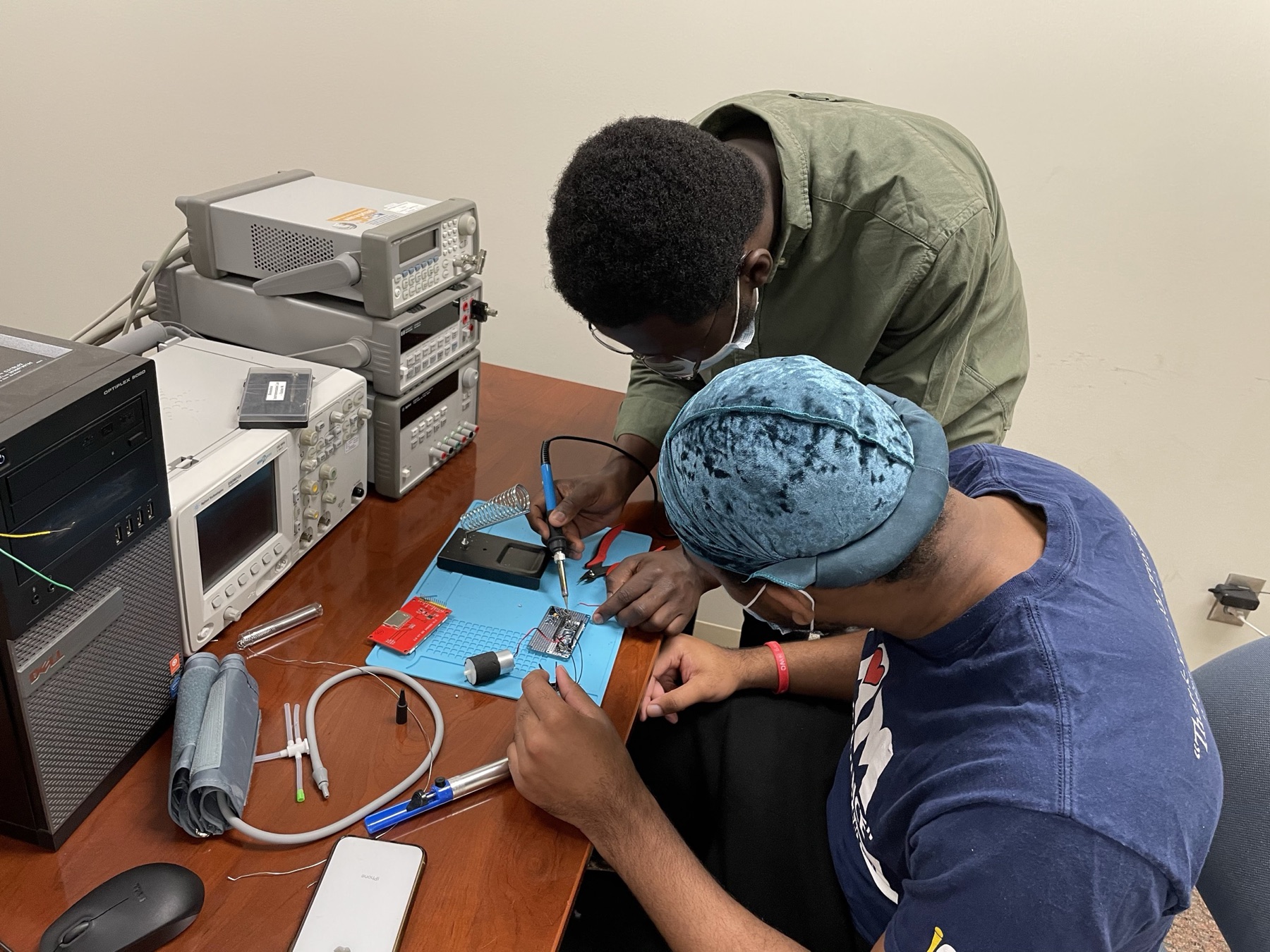 An instructor guiding a student through a soldering step at a bench with a signal generator and oscilloscope