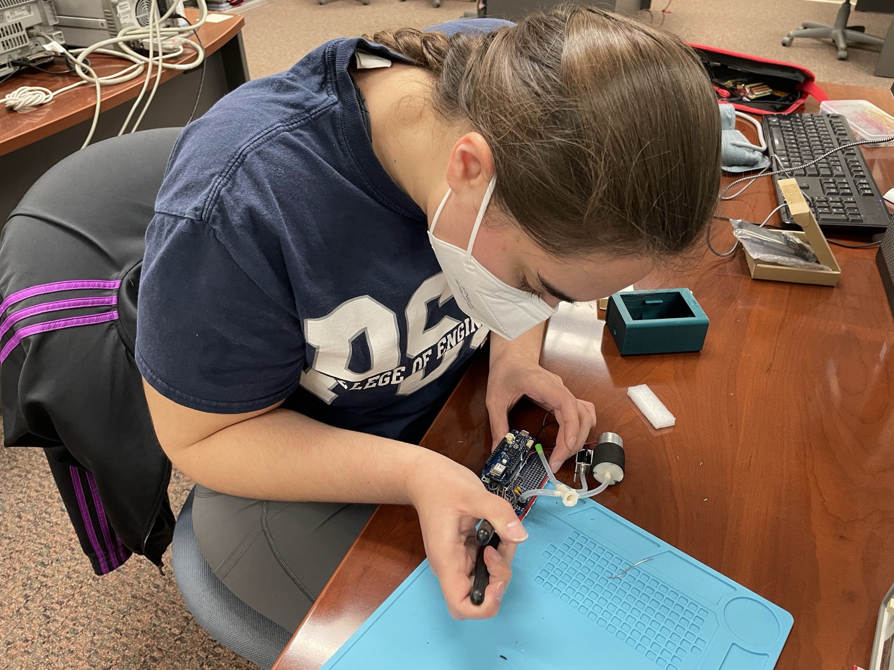 A student soldering the blood pressure monitor board, with the pump and tubing on the bench