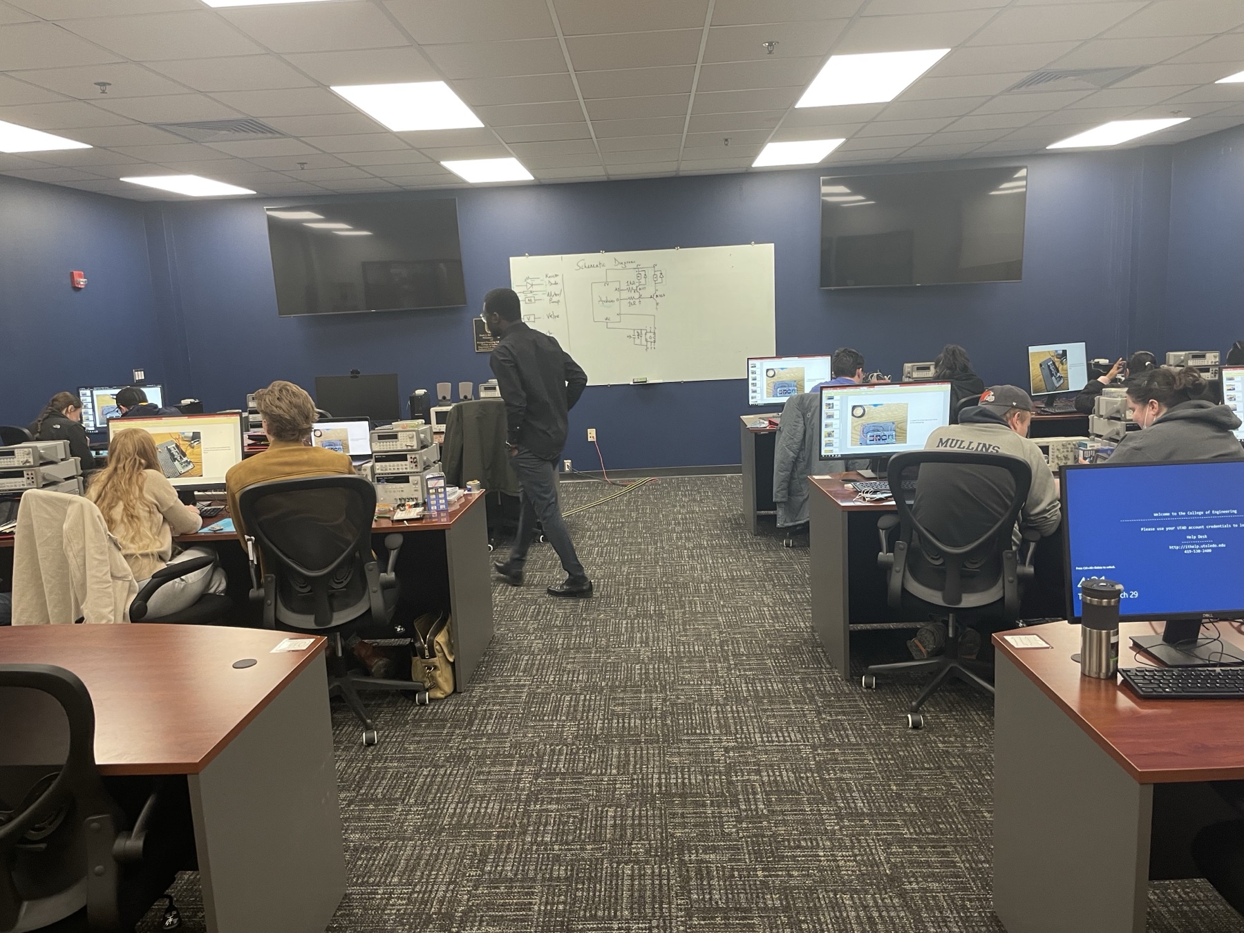 Solomon walking between student bench stations during a Medical Internet of Things lab session, with a circuit schematic on the whiteboard behind him